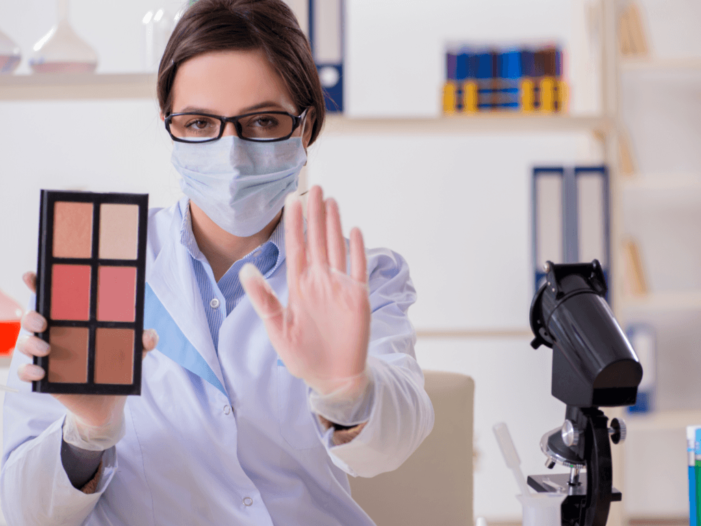 Seorang ahli kosmetik wanita mengenakan masker dan sarung tangan, memegang palet makeup, serta menunjukkan proses pengujian produk kosmetik di laboratorium